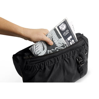 Bellroy Cinch Messenger Bag in black recycled nylon with hand placing newspaper inside front pocket