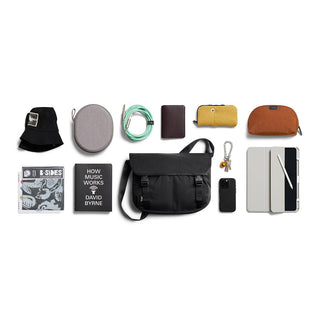 Bellroy Cinch Messenger Bag in black nylon with front buckles and adjustable shoulder strap displayed with accessories