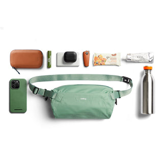 Bellroy Lite Sling crossbody bag in sage green made from recycled materials with multiple everyday items arranged nearby