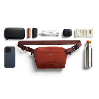 Bellroy Lite Sling Mini in rust red with adjustable strap and minimalist design next to everyday essentials