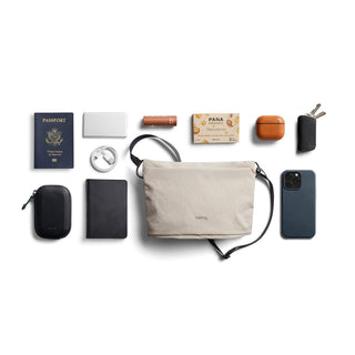 Bellroy Lite Sacoche in beige with travel essentials including phone, passport, keys, and portable charger arranged around it