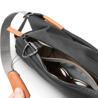 Bellroy Sling - 7L in black woven fabric with tan leather accents and zippered compartment holding sunglasses