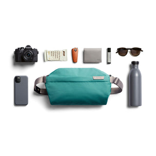 Bellroy Sling - 7L in teal with durable fabric, surrounded by everyday essentials and tech accessories
