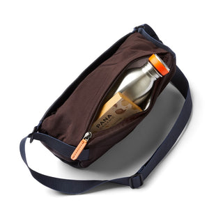 Bellroy Sling Mini Premium - 7L brown fabric sling bag with adjustable navy strap and partially open zipper