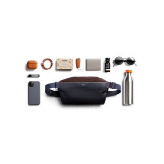 Bellroy Sling Mini Premium - 7L in navy and brown with everyday essentials arranged around it on white background