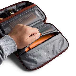 Hand placing an item inside waterproof compartment of Bellroy Hanging Toiletry Kit in brown tone