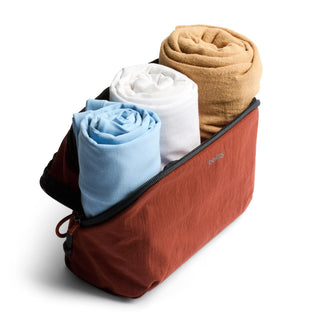 Bellroy Lite Packing Pouch 3L in rust color holding rolled clothing in blue, white, and tan fabrics