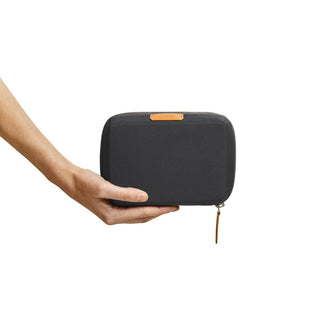 Hand holding a black Bellroy Tech Kit Compact with a tan zipper pull and top label against white background