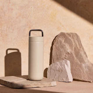 FELLOW Fellow Carter Carry Tumbler (20oz/591ml) in beige with stainless steel handle on stone surface