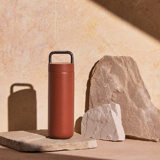 FELLOW Fellow Carter Carry Tumbler (20oz/591ml) in matte brown with stainless steel handle standing on stone slabs