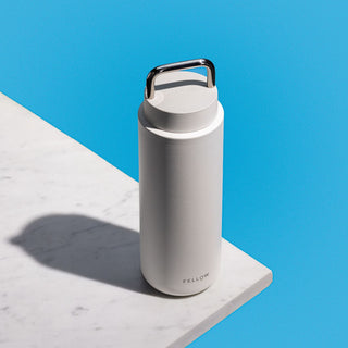 Fellow Carter Carry Tumbler (32oz/946ml) in white with stainless steel handle on marble surface against blue background