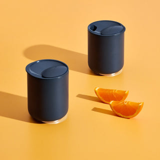 Two matte black FELLOW Rocky Lowball Tumblers (10oz/296ml) with lids on orange surface next to orange slices