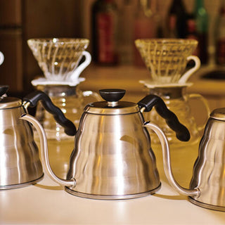 HARIO Buono Kettle 1200ml stainless steel with black handle, positioned on a kitchen counter with coffee drippers behind