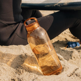 KINTO WATER Bottle 950ml in transparent amber copolyester with silicone handle resting on sand outdoors