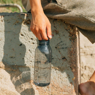 KINTO WATER Bottle 950ml held by hand, transparent copolyester body with black cap near concrete wall outdoors