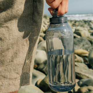 KINTO WATER Bottle 950ml held by hand, clear copolyester bottle filled with water outdoors by rocks