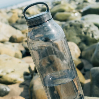 KINTO WATER Bottle 950ml transparent copolyester tumbler with black lid resting on rocky surface outdoors