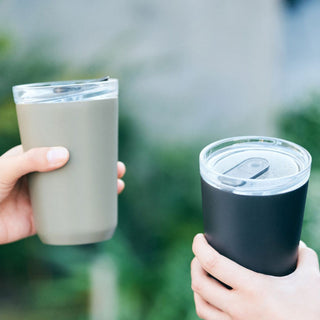 Two hands holding KINTO To Go Tumbler (with plug) 360ml in beige and black stainless steel design outdoors