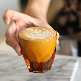 notNeutral VERO Cappuccino Glass 6oz held at an angle with latte art in amber tinted glass