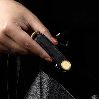 Orbitkey Saffiano Leather Key Organiser in black with gold-tone hardware held against black leather bag