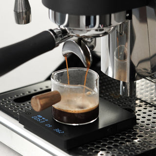 Timemore Black Mirror Mini Coffee Scale with a glass cup measuring espresso shot on a stainless steel machine