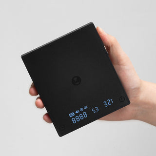 Hand holding Timemore Black Mirror Mini Coffee Scale with illuminated digital display on black surface