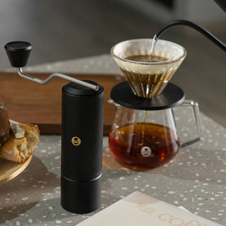 Timemore Chestnut X Lite hand grinder in black stainless steel beside pour-over coffee setup