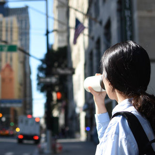 Woman drinking from white KINTO Travel Tumbler 500ml in urban setting with sunlight and buildings background